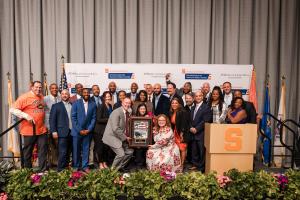 Group of 26 standing on platform and looking at camera, three people in front holding framed plaque