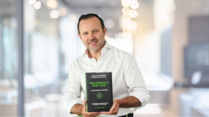 Stuart Lacey, stands confidently in an office environment, smiling at the camera and holding a book titled 'The Formula for Luck'. He is dressed in a casual white shirt, and the background is softly blurred with a warm light filtering through, highlightin