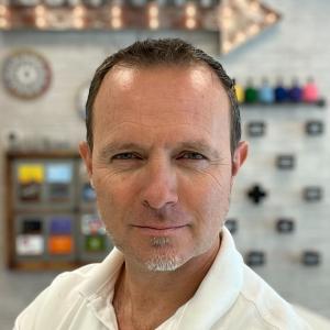 Close-up portrait of a man with a confident gaze, identified as Stuart Lacey. He has short, dark hair and is wearing a white shirt. The background is a creatively designed wall with various colorful elements, including a clock and a series of drawers, all