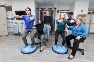 Two students balancing on a bosu ball holding dumbells while instructors correct their form.  Two additional students look on.