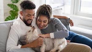 Two people sit on a chair snuggling with a happy white dog.
