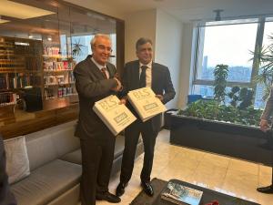Two men are standing in an office environment, each holding a folder with the BLS International & iDATA logos, symbolizing a formal business agreement. They are both dressed in business suits and smiling, suggesting a successful negotiation or partnership