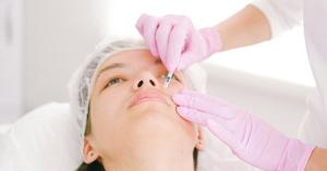 Un paciente recibiendo un procedimiento cosmético, mientras un profesional médico con guantes rosados administra cuidadosamente el tratamiento en el área del labio superior. El entorno sugiere un ambiente clínico con enfoque en la estética facial.