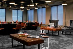 Modern meeting space at Moxy Phoenix Tempe/ASU with stylish furniture including leather chairs, a large central table with books, ambient lighting, and 'GOOD VIBES' pillow, set against a backdrop of large windows with shades.
