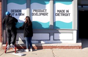 Two people putting up signs on the TUKAcenter that read "Design Cut & Sew," "Product Development," and "Made in Detroit"