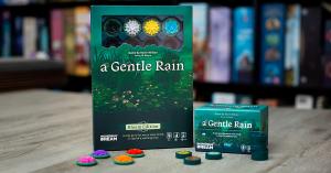 On the right, the remastered version of A Gentle Rain in its recognizable compact size. On the left, a tall box of A Gentle Rain (Bloom Edition) with a window showing some 3D sculpted flowers inside. Lily tokens are arranged next to these boxes on a table