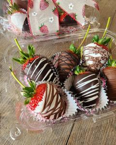 Six chocolate covered strawberries with drizzled chocolate as decoration. There are milk chocolate, white cholcolate, and dark chocolate covered berries. Each berry is in a paper cupcake-style cup and all six are in an open clear plastic box. In the backg