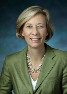 Executive headshot of Dr. Charlotte Sumner, a female with short blonde hair, wearing a green blazer, and against a blue background.