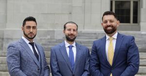 A photograph of three men wearing formal suits in shades of blue, with beards on their faces, standing in front of the courthouse steps in Miami, at the main entrance