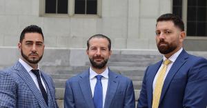 A photograph of three men wearing formal suits in shades of blue, with serious expressions and beards on their faces, standing in front of the courthouse steps in Miami, at the main entrance.