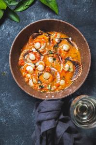 A red sauce with scallops and vegetables in a brown bowl next to a glass of water