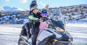 A tour guide with a helmet on, lifted above his head smiles with a child in the front seat of a Ski -Doo snowmobile. Mountains in the distance are snow covered with some shrubs in the light.
