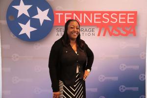 Smiling woman wearing a black dress stands next to Tennessee Self Storage association sign.
