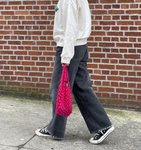 An image of someone that starts at their shoulders, holding a pink shibori bag. They're wearing a white crewneck sweater, black jeans, and black white top sneakers.