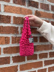 An arm with a white sleeve holding a pink shibori bag, small with a ruffled texture, in front of a brick background.