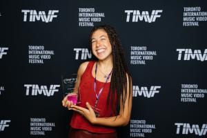 In the photograph, TYA, the winning artist of Best Music Video at the Toronto International Music Video Festival 2023, stands confidently in front of a step and repeat banner with the festival's logo. TYA is holding a trophy and smiling.
