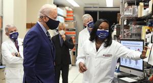 Dr. Kizzmekia Corbett, a young black woman dressed in a white lab coat shows her laboratory to President Joe Biden, a white man in his late seventies, who is dressed in a dark blue suit. Both are wearing masks.