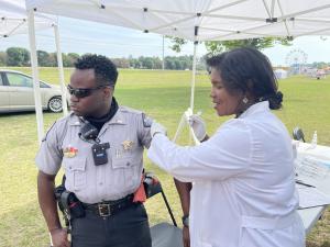 Dr. Karen Smith, a middle aged Black woman dress in a white doctor's jacket, administers the COVID-19 vaccine to a Black male police officer in uniform.
