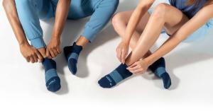 A man and a woman sit cross legged on the ground wearing blue running clothes and navy wicked comfort socks.