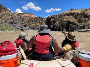 Three BIPOC guests sit at the front of a raft in the middle of a muddy river. One of the guests holds the other guest's PFD and points to the scenery depicting black schist rock canyon walls.