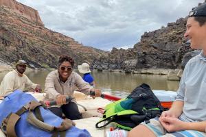 Three BIPOC guests are on a raft while one of them rows the raft. The guide sits in the corner and looks on while the guest practices rowing.