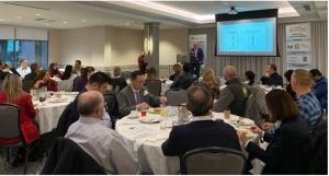 Andy Kovacs, President of Moments of Truth Insurance Services, stands at a podium presenting to an audience at the Markham Board of Trade Breakfast Networking Event. He is gesturing towards a projection screen displaying a presentation slide, with attende