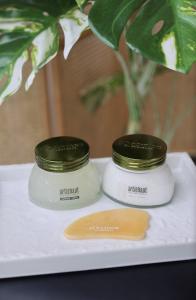 A glass jar of body scrub next to a glass jar of white body cream, behind a Gua Sha on a white tray