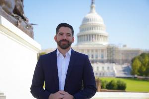 Photo of a smiling Luis Dávila Pernas from the waist up, wearing a white shirt and dark blue suit jacket, arms crossed, standing in front of the US Capitol Building in Washington, DC