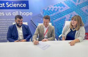 Three people sat at a table with one signing a contract. The other two are smiling