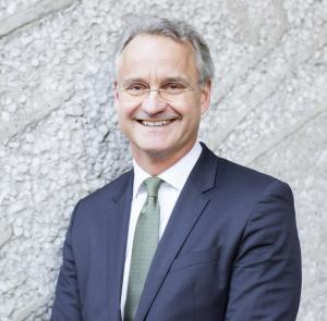 The photo features Magnus Billing, senior advisor at RS Metrics. He is wearing glasses and a navy blue suit with a white button-up shirt and a green tie. He is standing in front of a light gray wall and smiling.