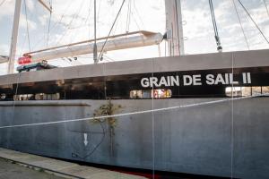The side of a gray and black boat with the words "Grain de Sail II" on it, with a wine bottle being smashed against it