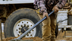 A construction worker pours cement for a sidewalk project as part of concrete works, and a mixer truck is shown behind him.