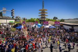 Large group of people in San Francisco, Japantown gathered to watch a parade. The parade is featuring a mikoshi (shrine) that is being carried by dozens of people--many in cultural attire who are celebrating the cherry blossom festival