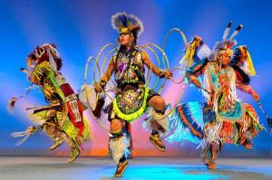 Three Native American men perform on stage in full regalia