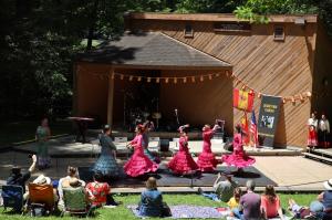 Female dancers wearing colorful long dresses while twirling around on an amphitheater stage surrounded by trees and audience sitting on the grass.
