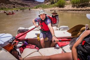 A man with a PFD on exlaims while using both hands to row an 18 foot raft with two oars. Another guest sits on the raft behind him and watches.