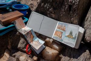 A sketch journal shows a watercolor painting of orange canyon walls and a blue river with some green trees. In the background of the image, a cup of water and a wooden box with watercolor tools are visible. These items are staged on dark rock in the wilde