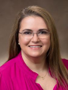 Mindy Beckwith, RN brunette smiles wearing glasses and a pink shirt on brown background