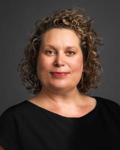 Mariah Chrans smiles in her headshot against a grey background. She has blonde curly hair and a black dress. She is smart and business minded.