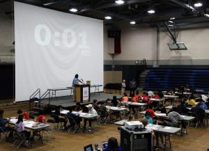 Students work at tables in a high school gym as a large projector displays "0:01"