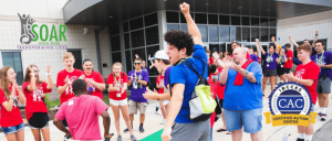 A variety of people wearing red, blue, violet, and pink shirts are clapping their hands and celebrating.