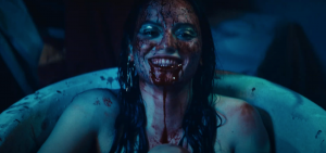 Singer Xaya laughs hysterically while sitting in a bathtub filled with red liquid. She holds a cup to her lips, appearing to drink from the tub. The liquid obscures her entire naked body, except for her arms and shoulders.