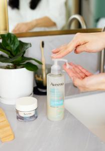 A small white-capped jar of cream-colored moisturizer next to a pump bottle of cleanser on a marble gray bathroom sink counter, with a woman's hand pumping the bottle into her palm