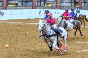 4 polo players ride horseback in enclosed polo arena with umpire following