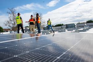 Men in bright work vests and hard hats talk with solar panels in the foreground.