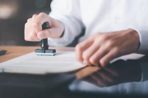 The image depicts a businessman validating and managing business documents and agreements. He is shown signing a business contract, indicating approval of contract documents with a confirmation or warranty certificate. An approval stamp is visible on the