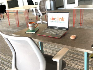 Gray chair pushed into a gray chair with a laptop, mouse pad and mouse, cup of coffee on top. The computer screen reads "the link."