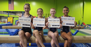 Four ILA student gymnasts sit on a balance beam holding 'first day of school' signs.