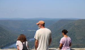 Two tween girls stand on either side of a middle aged man overlooking the valley and river below as they lean on a stone wall.