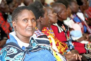 A woman in Kenya looks to camera with a serious expression, behind her are rows of women sitting down and looking in the other direction. They are all wearing brightly colored clothes that are traditional to the Southern Rift Valley in Kenya.